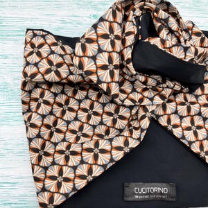 May include: A black and patterned scarf with a geometric floral design in shades of orange, gray, and black. The scarf is folded, revealing a solid black underside. A small label reads "CUCITORINO - Be yourself, love yourself."