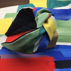 Op de afbeelding: Een kleurrijk geknoopt hoofdband met strepen in rood, geel, blauw, groen en wit. Het hoofdband is gemaakt van stof en heeft een zwart elastiek.