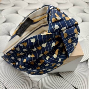 May include: A navy blue knotted headband with a repeating pattern of white and gold shapes and vertical lines. The interior lining features yellow and white stripes. The brand name "CUCTORINO" is visible on a black tag.