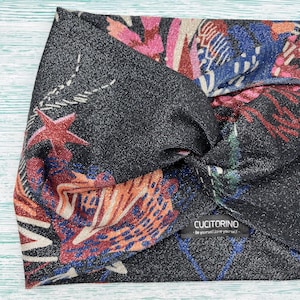 Op de afbeelding: Een zwarte, glinsterende hoofdband met een kleurrijk bloemen- en sterrenpatroon. Het ontwerp bevat rode, blauwe, roze en oranje elementen. De hoofdband heeft een gedraaide knoop en een klein label met de tekst "CUCITORINO - Be yourself! Love yourself!"