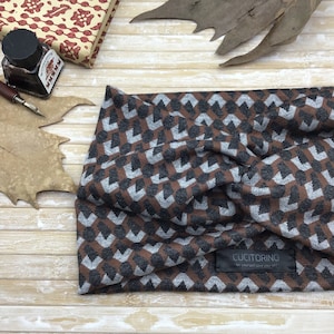 May include: A brown, gray, and silver geometric patterned headband. The headband is made of a soft fabric and features the brand name "CUCITORINO" on a black label. The headband is displayed on a light wood surface.