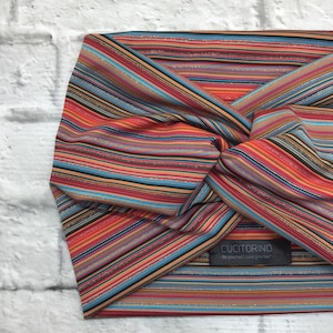 Può includere: Un turbante per capelli a righe colorate con un luccichio argentato. Il turbante è realizzato in un tessuto morbido ed elastico e ha un design annodato. Il turbante è etichettato "CuciTorino - Sii te stesso, amati!"