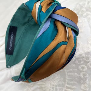 Può includere: Una fascia annodata con un motivo geometrico turchese, marrone e blu. La fascia ha una fascia larga in tessuto verde acqua unito e un'etichetta con la scritta "CUCITORINO". Questo accessorio è adatto a vari tipi di acconciatura.