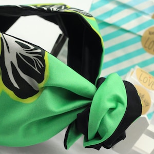 May include: A green and black fabric headband with a leaf pattern. The headband has a knotted design and is made of a silky material. The background includes gift bags and tags.