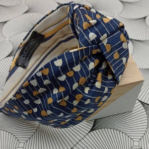May include: A navy blue knotted headband with a repeating pattern of white and gold half-circles and vertical lines. The interior lining features yellow and white stripes. The label reads "CUCITORINO - be yourself, everyday!"