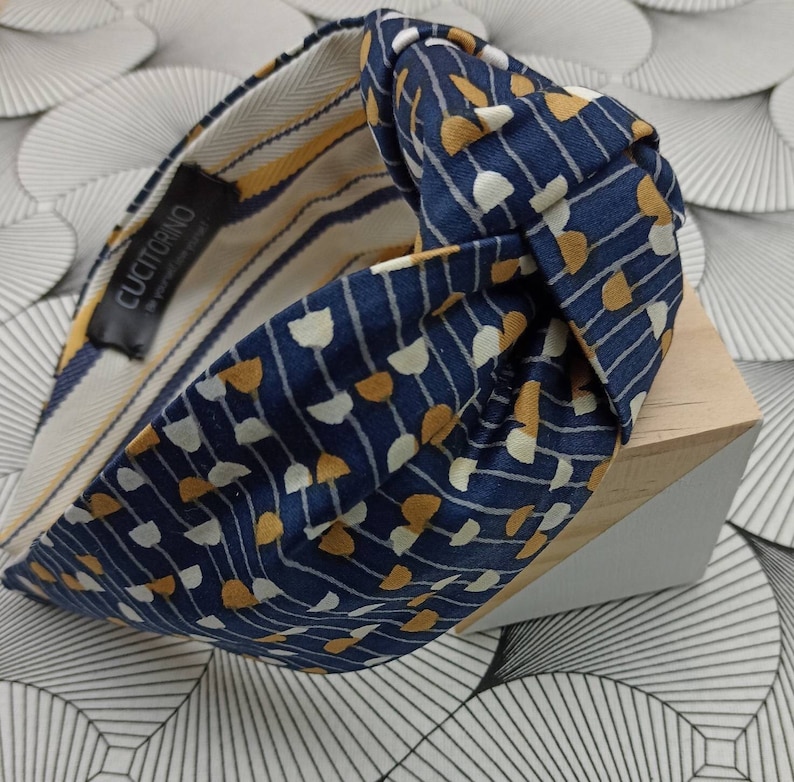 May include: A navy blue knotted headband with a repeating pattern of white and gold half-circles and vertical lines. The interior lining features yellow and white stripes. The label reads "CUCITORINO - be yourself, everyday!"