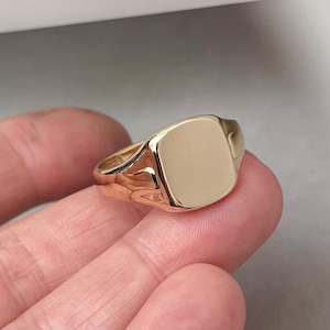 Könnte beinhalten: Ein goldener Siegelring mit quadratischer Oberfläche. Der Ring hat eine warme, polierte Goldfarbe und ein klassisches, elegantes Design. Der Ring wird an einem Finger gezeigt.