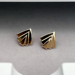 May include: A pair of gold-toned stud earrings featuring a fan-like shape with three distinct, layered triangular segments. The metal has a polished, reflective surface that catches the light, creating a smooth, geometric appearance.