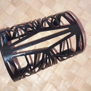 Może przedstawiać: Czarny metalowy cylindryczny uchwyt z geometrycznym ażurowym wzorem. Uchwyt ma szereg trójkątnych i liniowych wycięć. Obiekt spoczywa na jasnobrązowej tkanej powierzchni.