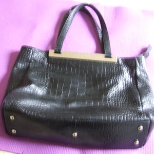 May include: A black handbag with a textured crocodile-embossed pattern, featuring two top handles, a metallic silver-toned bar accent at the top center, and four small silver-toned metal feet on the bottom base for stability and protection.