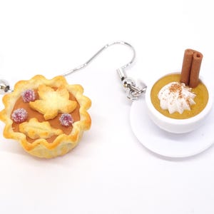 May include: A pair of novelty earrings featuring miniature food items. One earring is a pie with a leaf design and berries. The other is a small bowl of pumpkin pudding with whipped cream and a cinnamon stick.