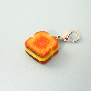 May include: A miniature charm shaped like a grilled cheese sandwich. The charm features two slices of orange-brown bread with a yellow cheese filling. It has a silver-colored clasp for attaching to a bracelet or necklace.