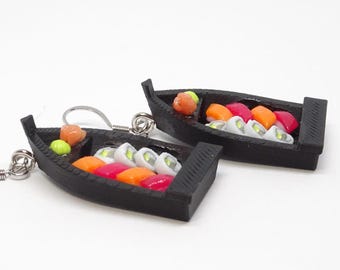 Könnte beinhalten: Ein Paar Sushi-Boot-Ohrringe. Jedes schwarze Boot ist mit farbenfrohen Sushi-Rollen gefüllt, darunter orange, rosa und weiße Sorten. Die Ohrringe sind vor einem weißen Hintergrund platziert.