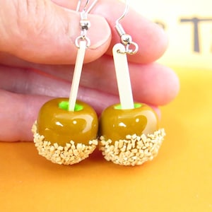 May include: A pair of dangle earrings featuring miniature caramel apple charms with a glossy brown coating, a bright green interior, and a textured base covered in small beige nut-like sprinkles, attached to silver-toned metal hooks.