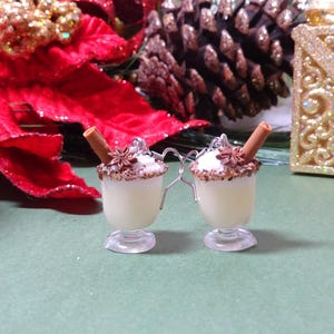 May include: Two miniature eggnog ornaments with cinnamon sticks and star anise. The eggnog is in clear, stemmed glasses, topped with whipped cream and a brown sugar rim. Silver wire connects the two ornaments. Festive holiday decor.