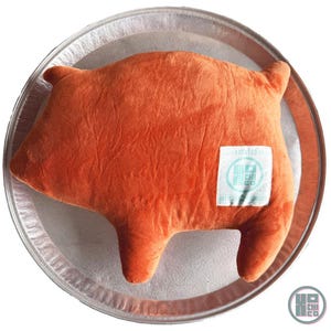 May include: An orange pig-shaped plush pillow with a textured surface, resting on a silver tray. The pillow has a small white tag with blue text and a logo. The pillow is a fun and whimsical decorative item.