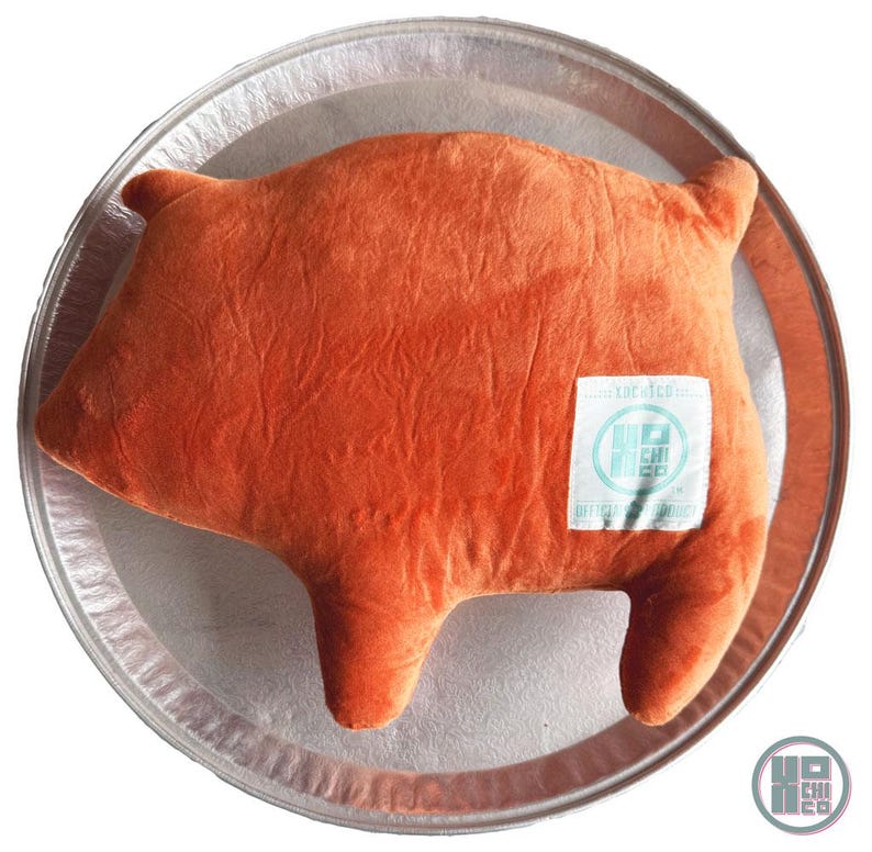 May include: An orange pig-shaped plush pillow with a textured surface, resting on a silver tray. The pillow has a small white tag with blue text and a logo. The pillow is a fun and whimsical decorative item.
