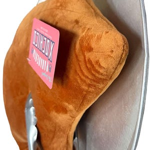 May include: A plush, brown, food-shaped pillow resembling a piece of fried chicken, held by metal tongs, and placed on a silver platter. A pink tag with white text is attached to the pillow. The image has a white background.