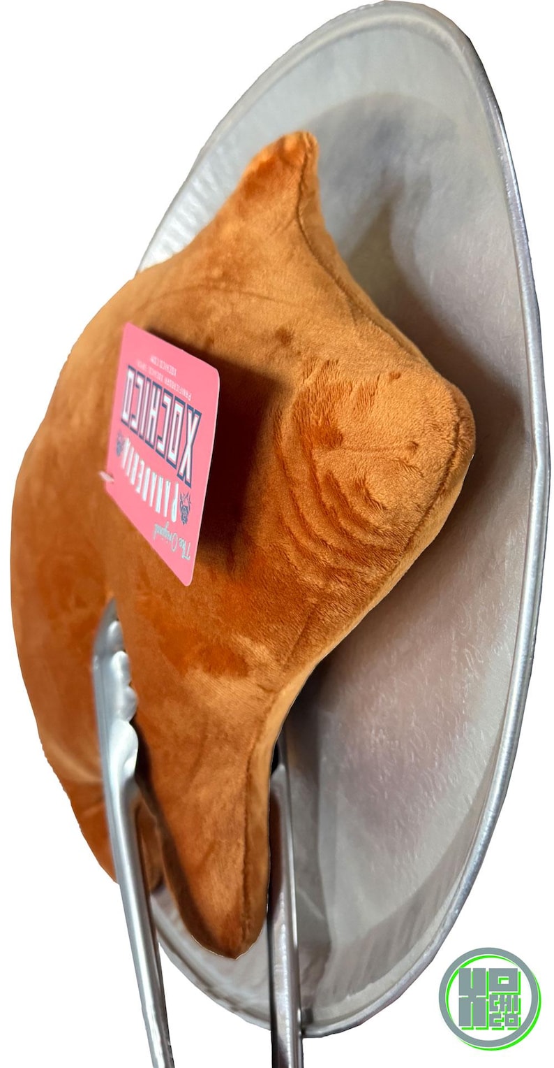 May include: A plush, brown, food-shaped pillow resembling a piece of fried chicken, held by metal tongs, and placed on a silver platter. A pink tag with white text is attached to the pillow. The image has a white background.