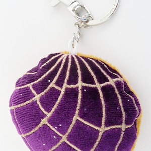 May include: Purple and gold plush keychain shaped like a concha, a traditional Mexican sweet bread. The keychain has a silver metal ring and clasp.