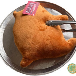 May include: A plush, orange-brown stuffed toy shaped like a cooked chicken, resting on a silver platter. Metal tongs are gripping the toy. A pink tag with white text is attached to the toy. The toy is a humorous take on a cooked chicken.