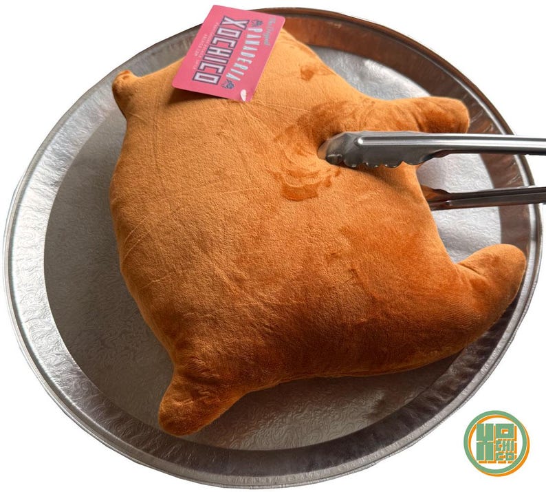 May include: A plush, orange-brown stuffed toy shaped like a cooked chicken, resting on a silver platter. Metal tongs are gripping the toy. A pink tag with white text is attached to the toy. The toy is a humorous take on a cooked chicken.