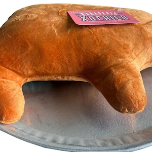May include: A plush, brown croissant-shaped pillow rests on a silver tray. A pink tag with the text "PANADERIA XOCHICO" is attached to the pillow. The pillow is soft and textured, resembling a baked pastry.