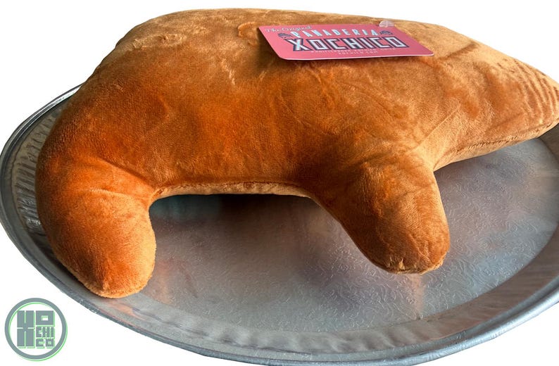 May include: A plush, brown croissant-shaped pillow rests on a silver tray. A pink tag with the text "PANADERIA XOCHICO" is attached to the pillow. The pillow is soft and textured, resembling a baked pastry.