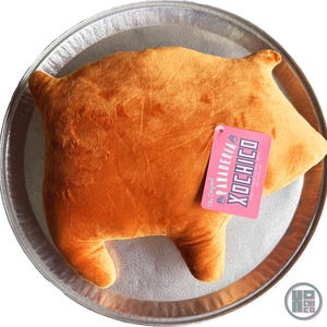 May include: A plush, orange-colored pillow shaped like a pig, resting on a silver tray. The pillow has a tag that reads "Panaderia Xochico" in pink and blue. The pillow is a fun, whimsical item.