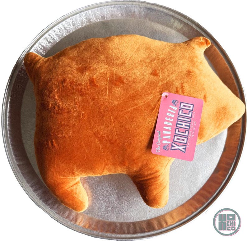 May include: A plush, orange-colored pillow shaped like a pig, resting on a silver tray. The pillow has a tag that reads "Panaderia Xochico" in pink and blue. The pillow is a fun, whimsical item.