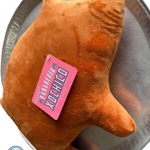 May include: A plush, orange-brown bear-shaped pillow with a tag that reads "PANADERIA XOCHICO". The pillow is resting on a silver-colored, decorative tray. The tag is pink with white and blue text.