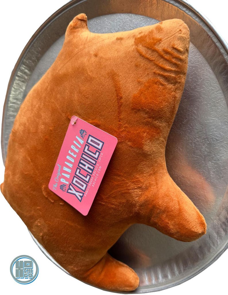 May include: A plush, orange-brown bear-shaped pillow with a tag that reads "PANADERIA XOCHICO". The pillow is resting on a silver-colored, decorative tray. The tag is pink with white and blue text.