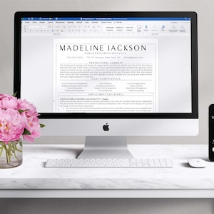 May include: A white desk with a silver iMac computer displaying a resume for Madeline Jackson, Human Resources Executive. The resume includes a professional summary, core competencies, and employment experience. A white keyboard and a black smartphone with the text "Your Dream Job" are also on the desk. A vase of pink peonies sits on the desk in the foreground.