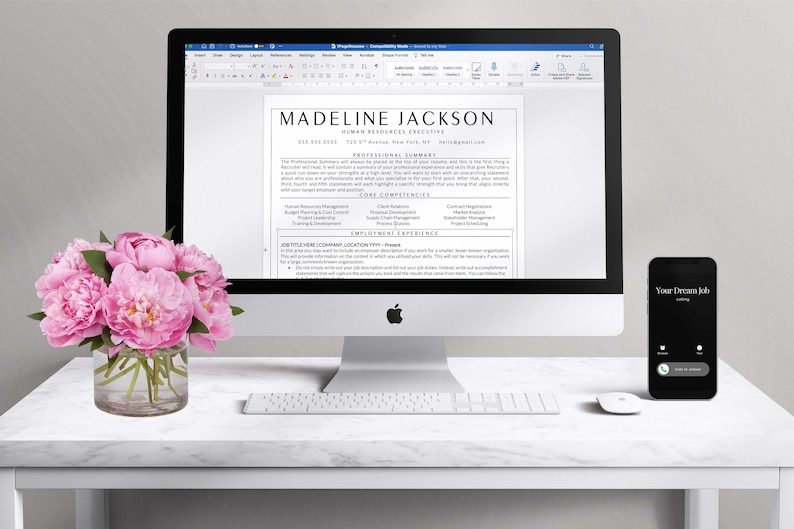 May include: A white desk with a silver iMac computer displaying a resume for Madeline Jackson, Human Resources Executive. The resume includes a professional summary, core competencies, and employment experience. A white keyboard and a black smartphone with the text "Your Dream Job" are also on the desk. A vase of pink peonies sits on the desk in the foreground.