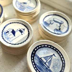 Op de afbeelding: Stapels kleine, ronde, witte keramische borden met blauwe nautische illustraties. Ontwerpen omvatten een vuurtoren, een zeilboot en een zeeman. Elk bord heeft een blauwe rand.