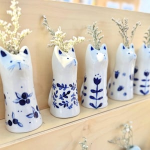 May include: A collection of white ceramic cat-shaped vases with blue painted details and floral designs. Each vase holds dried white flowers. The vases are arranged on a light wooden shelf.