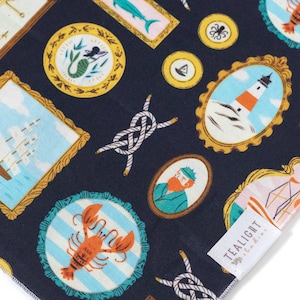 May include: A navy blue fabric with a repeating pattern of framed illustrations. The illustrations include a ship, a lighthouse, a mermaid, a lobster, a sailor, an anchor, and a knot. The fabric has a tealight label with the text "Tealight" and a small logo.