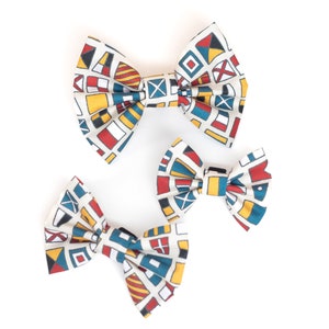 May include: Three fabric bow ties with a nautical flag pattern in shades of blue, red, yellow, and white. The bow ties are arranged in a triangular shape on a white background.