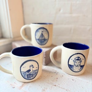 Puede incluir: Tres tazas de cerámica con exteriores color crema e interiores azul marino. Cada taza presenta una ilustración de temática náutica en azul marino: un velero, una gaviota y un perro con gorro de capitán.