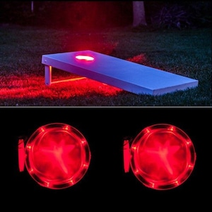 May include: Red LED lights illuminate a white cornhole board and two red LED disc lights. The lights are on a dark background.