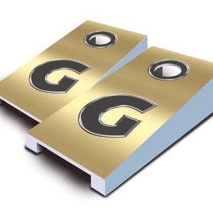 May include: Two gold-colored cornhole boards with black letter G's on each board. The boards have a white base and a black circle in the center.
