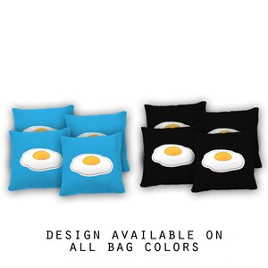May include: Six bean bags with a design of a fried egg on them. Three bean bags are blue and three are black. The text "DESIGN AVAILABLE ON ALL BAG COLORS" is below the bean bags.