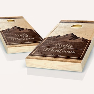 May include: Two wooden cornhole boards with a mountain landscape and the text "Carly and Montana" in a brown and gold frame. The boards are made of natural wood and have a rustic look.