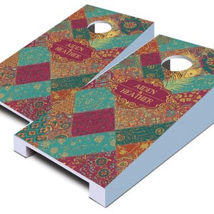 May include: Two cornhole boards with a colorful patchwork design in teal, burgundy, and gold. Each board has a circular hole and the text "AIDEN & HEATHER" in a decorative frame. The boards have light blue sides.