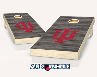 Puede incluir: Un juego de dos tablas de cornhole de madera con un acabado gris desgastado. Las tablas presentan un logotipo rojo de la Universidad de Indiana con las letras "IU" y un símbolo de marca registrada. Las tablas están preparadas para un juego de cornhole.