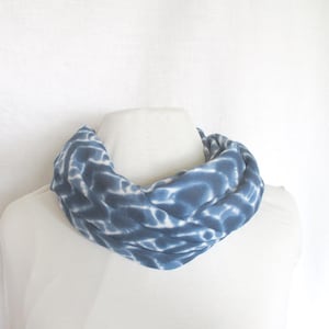 May include: A blue and white tie-dye infinity scarf. The soft, circular scarf is displayed on a white mannequin. The fabric has a pattern of blue swirls and white accents. This accessory is a stylish addition to any wardrobe.