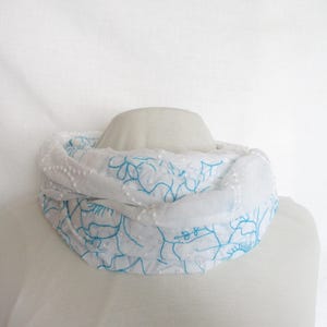 May include: White infinity scarf with blue floral embroidery. The scarf is made of a lightweight fabric and is displayed on a mannequin. The design features delicate blue lines and small white dots.