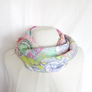 May include: A colorful floral patterned scarf in shades of pink, green, blue, and yellow. The scarf is folded into a circle and is displayed on a white surface.
