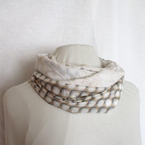 May include: A beige and white infinity scarf with a repeating pattern of grey and white shapes. The scarf is draped around a mannequin's neck, showcasing its design and texture. The scarf is made of a soft, lightweight fabric.