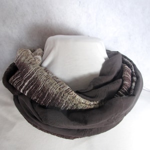 May include: A brown infinity scarf with a textured, patterned design. The scarf is wrapped around a white mannequin, showcasing its layered construction and color variations. The scarf's design includes shades of brown, beige, and white.